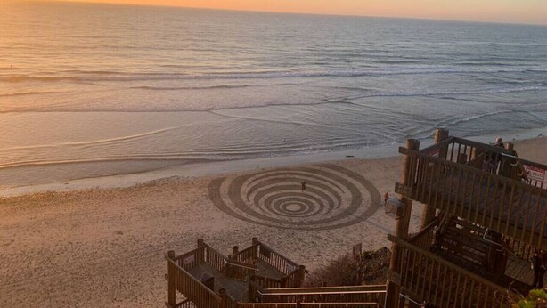 Ein Strand mit einem spiralförmigen Muster im Sand, eine Holztreppe, die zum Strand hinabführt, und ein Holzzaun im Vordergrund.