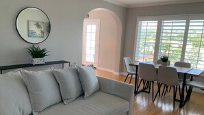 Ein modernes Wohnzimmer mit grauem Sofa, einem Esstisch mit Stühlen, einem großen Fenster mit Jalousien und einer Zimmerpflanze.