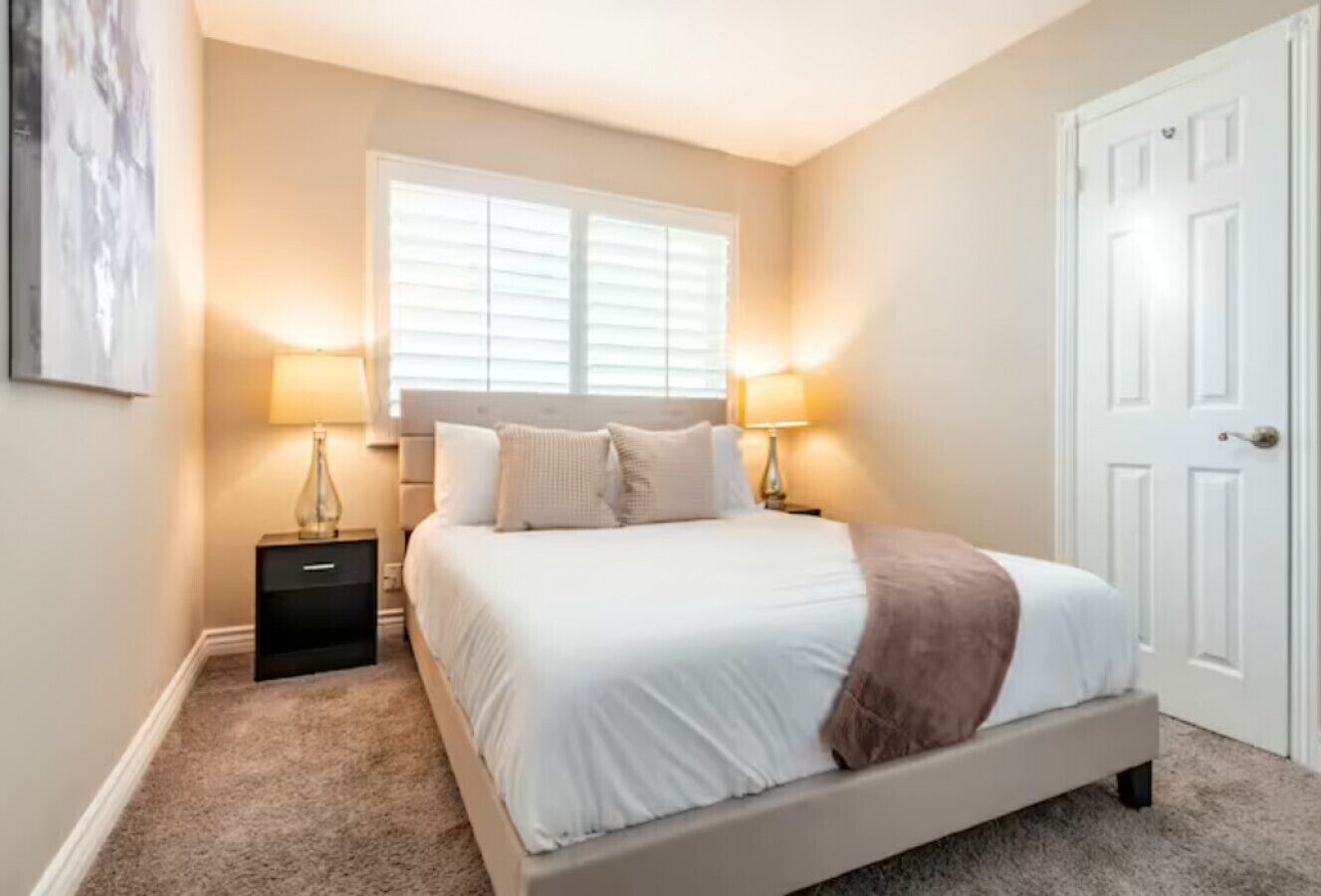 Un dormitorio con cama, mesitas de noche, una ventana y una puerta.