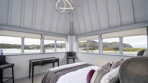 A bedroom with a bed, a desk, and two chairs. It has large windows with a view of the sea and hills.
