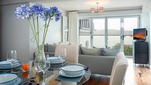 A modern dining area with a glass table set for four, a vase with blue flowers, and a view of the outdoors through large windows.