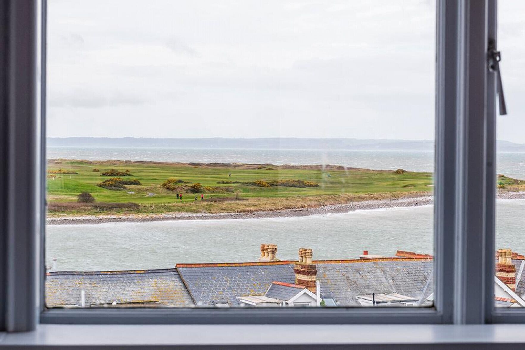 A coastal view from a window overlooking a beach, grassy fields, and a distant shoreline.