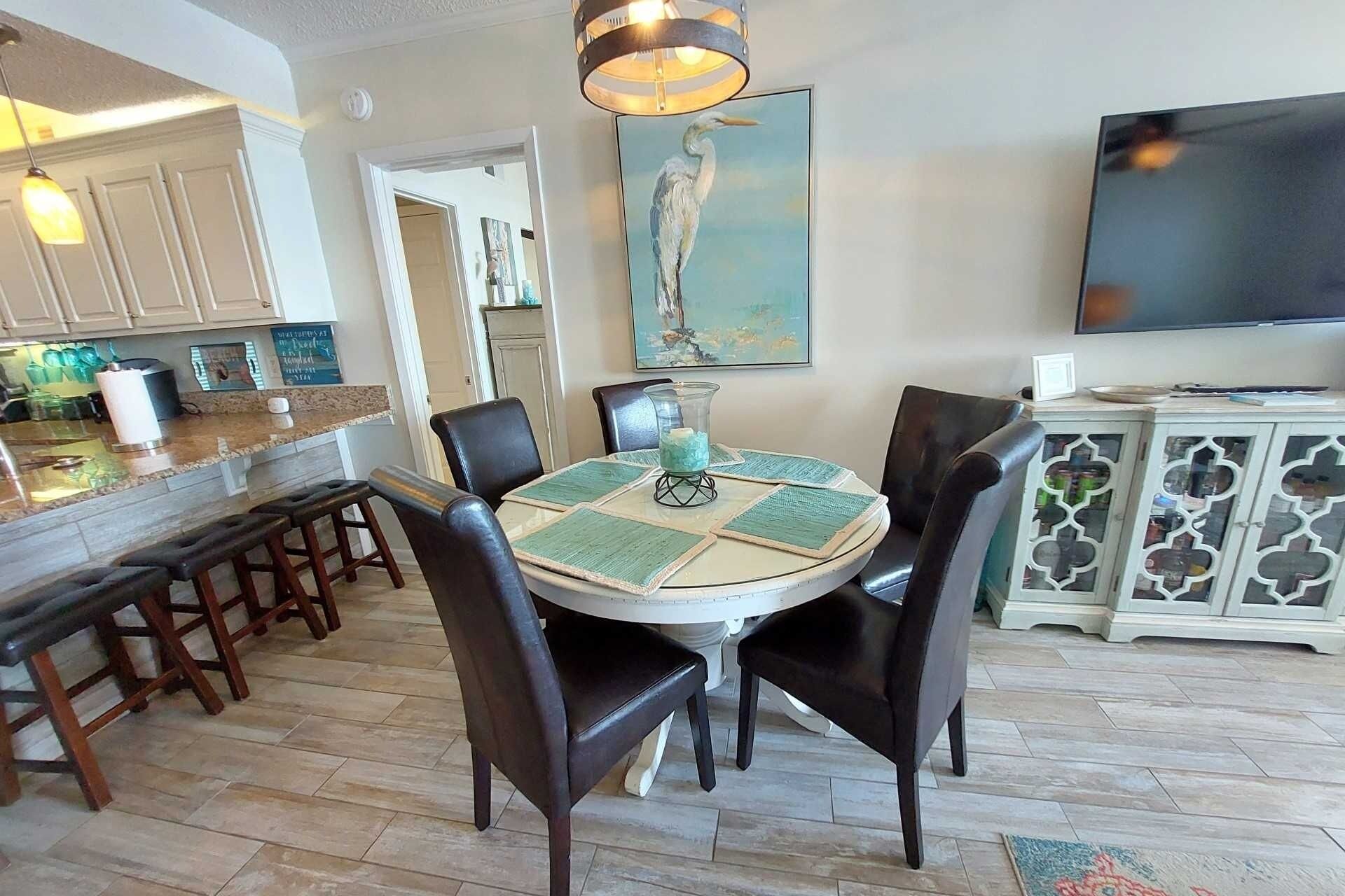 A dining area with a round table, chairs, a TV, and a kitchen in the background.