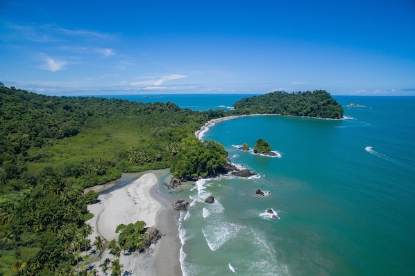 An aerial view of a coastal landscape with a beach, dense greenery, and clear blue waters.