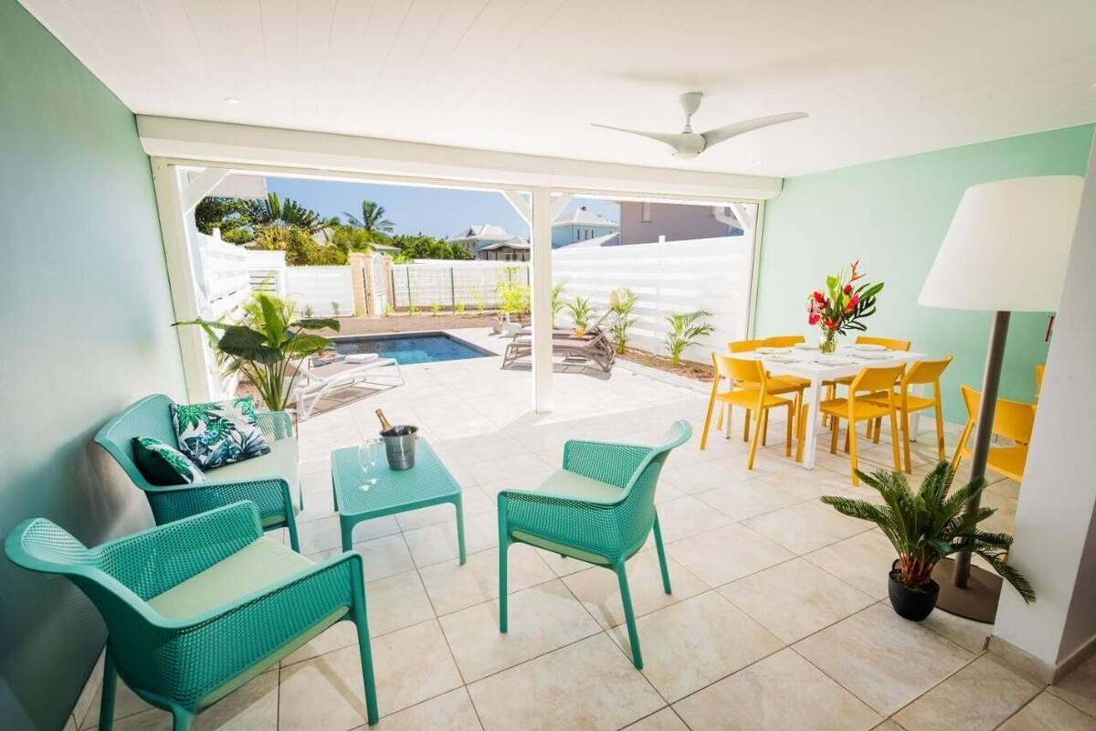 A bright room with a pool, green chairs, a table, and a ceiling fan.