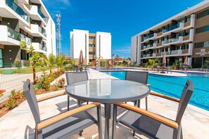 A poolside area with a round glass table and chairs, surrounded by a pool and modern buildings.