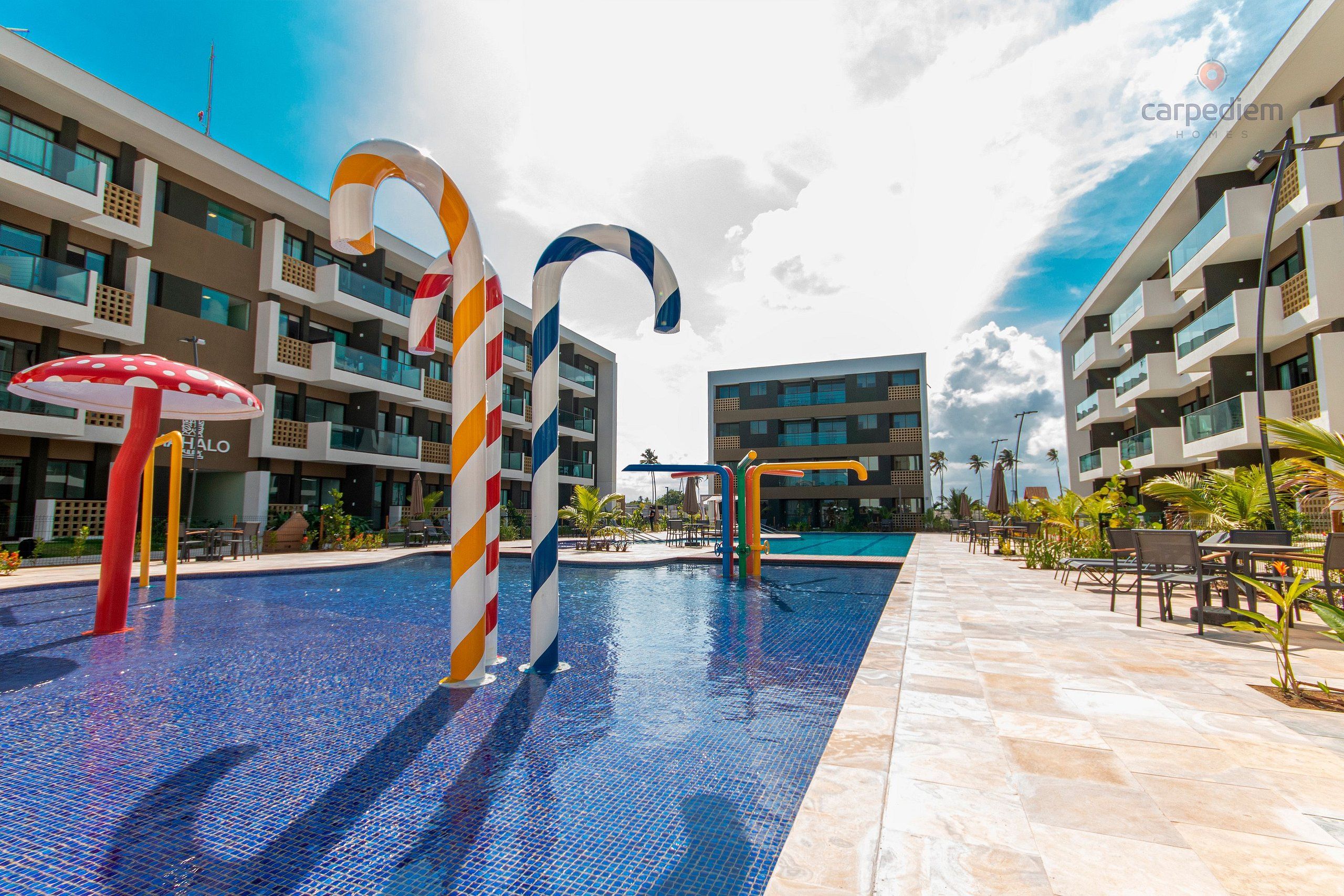 Área de piscina com decorações coloridas de cana de açúcar, uma escultura de cogumelo vermelho e um edifício moderno com varandas e piscina ao fundo.