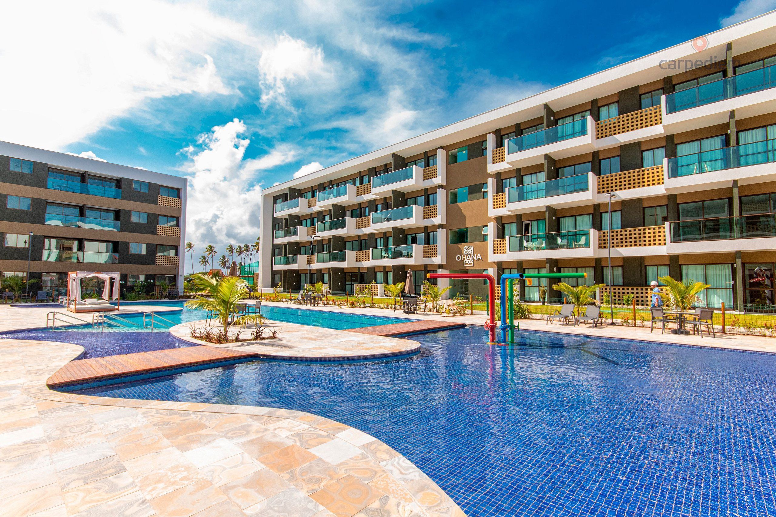 A modern hotel with a large outdoor swimming pool, multiple balconies, and a well-maintained garden area.