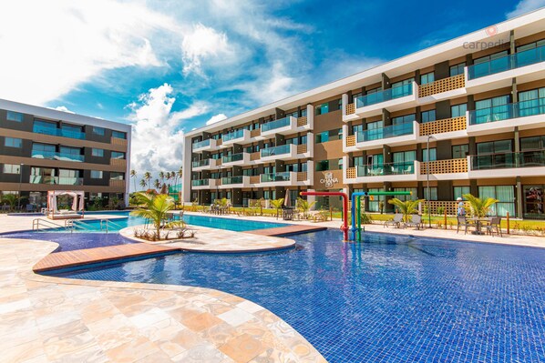 A modern hotel with a large outdoor swimming pool, multiple balconies, and a well-maintained garden area.