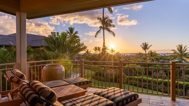 Un balcon doté de chaises longues et d’un grand vase, donnant sur un jardin aménagé et des palmiers, avec une vue imprenable sur le coucher de soleil.