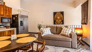 Un salon comprenant un canapé, une statue de Bouddha, une table à manger avec des sets de table, et un coin cuisine équipé d’appareils électroménagers.