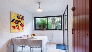 Een eetgedeelte met een witte tafel, stoelen, een plafondlamp, een raam met uitzicht op groen, en een deur.