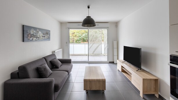 Ein modernes Wohnzimmer mit grauem Sofa, einem Holztisch, einem Flachbildfernseher auf einem Holzboden und einem großen Fenster mit Blick auf Grünflächen.