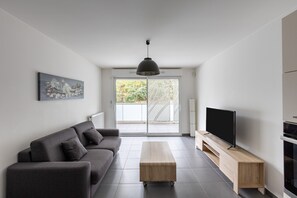 A modern living room with a grey sofa, a wooden coffee table, a flat-screen TV on a wooden stand, and a large window with a view of greenery. - Carré Huit - Two Bedroom Apartment, Sleeps 6 (Marseille)