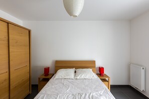A bedroom with a wooden wardrobe, a wooden nightstand with a red lamp, a white bed with two pillows, and a white radiator. - Carré Huit - Two Bedroom Apartment, Sleeps 6 (Marseille)