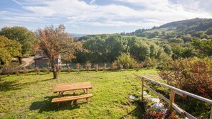 Ein Picknickplatz mit einer Holzbank und einem Geländer, umgeben von üppigem Grün und einem Steinhaus im Hintergrund.
