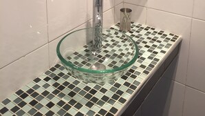 Ein Badezimmer mit einem Waschbecken aus Glas, einem runden Spiegel mit Mosaikfliesen, einer kleinen Ablage und einem Handtuchhalter.