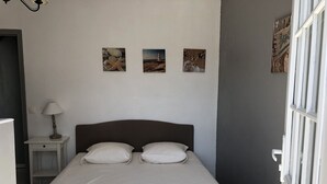 Ein Bett mit zwei Kissen, ein Nachttisch mit Lampe und drei Wandbilder, die einen Leuchtturm, Felsen und eine strukturierte Wand zeigen.