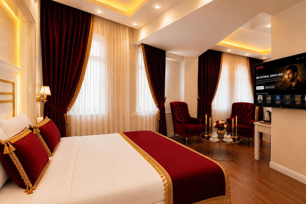 A hotel room with a large bed, two red armchairs, a small table with a candle, and a wall-mounted TV displaying a movie.