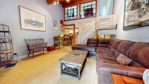 Ein geräumiges Wohnzimmer mit einem großen braunen Ledersofa, einem Holztisch und einer hölzernen Bank. Eine Treppe führt in einen oberen Bereich, und im Hintergrund ist eine Küchenzeile erkennbar.