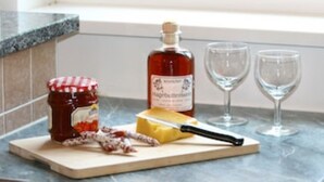 Un tagliere di legno con un vasetto di marmellata, una bottiglia di miele, un pezzo di formaggio, un coltello e due bicchieri da vino su un piano di lavoro in cucina.