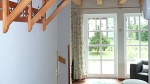 Una stanza con una scala in legno, un tappetino verde, una panca e vista su un giardino attraverso ampie finestre.