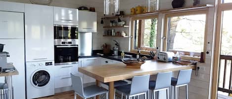 A compact kitchen with a wooden table, four chairs, a washing machine, and a window with a view of trees.