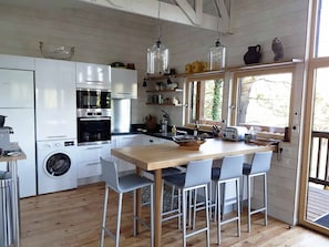 A compact kitchen with a wooden table, four chairs, a washing machine, and a window with a view of trees. - Le Chalet du Carla - SPA - Riverfront (Castelnau-de-Lévis)