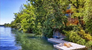 Un muelle se adentra en un lago tranquilo, rodeado de exuberante vegetación y un edificio de madera con balcón.