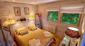 Un dormitorio con cama, mesita de noche con lámpara, ventana con vistas a la vegetación y estante con libros y objetos decorativos.