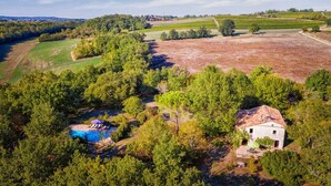 Une vue aérienne d’une propriété dotée d’une piscine entourée d’arbres, d’une petite maison avec un toit en tuiles et de vastes champs ouverts.