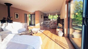 A spacious living area with a sofa, a coffee table, and a dining area with chairs. There is a large window providing a view of the outdoors.