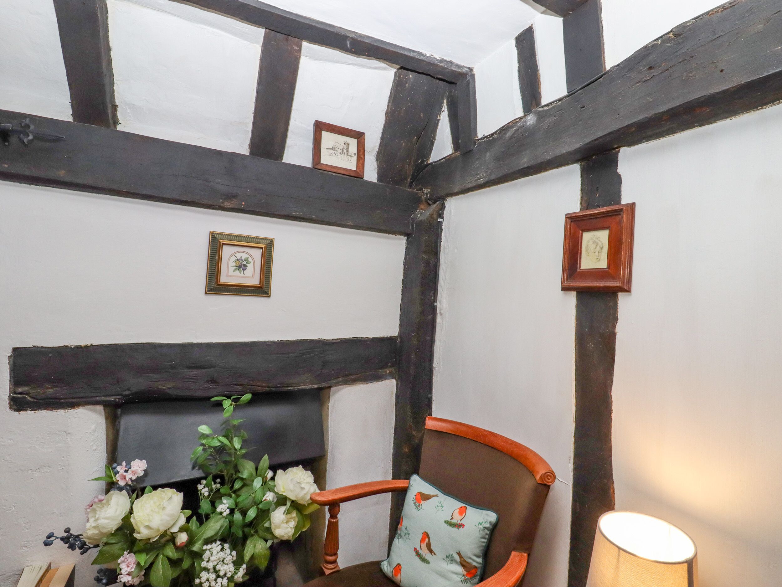 A room with exposed wooden beams, a fireplace, a chair with a floral cushion, and framed pictures on the wall.