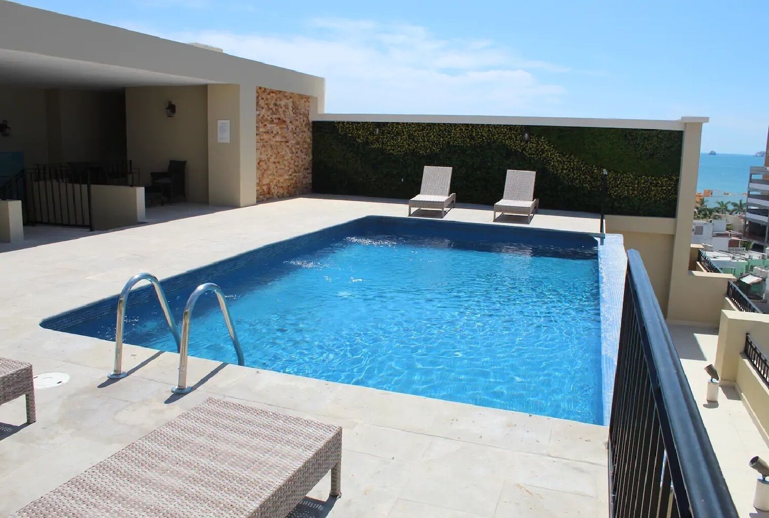 Une piscine rectangulaire avec un escalier en métal, entourée de chaises longues et d’un mur de pierre agrémenté de verdure.