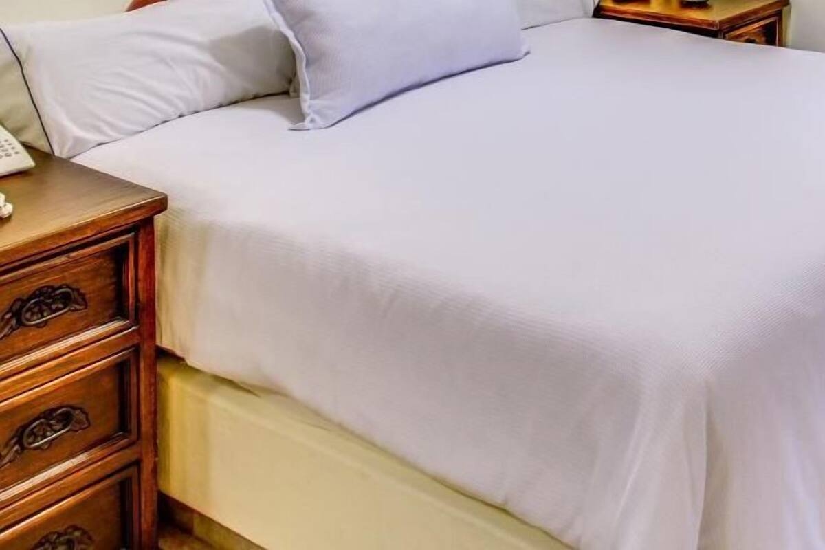Una cama bien hecha con un edredón blanco, una mesita de noche de madera con cajones y una mesita auxiliar con una lámpara.