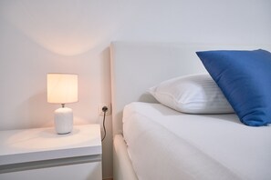 Una cama bien hecha con ropa blanca y una almohada azul, una mesita de noche blanca con una lámpara, y un enchufe en la pared.