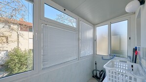 Un baño moderno y luminoso con paredes de azulejos blancos, amplias ventanas con persianas y un cesto de ropa blanca sobre una encimera.
