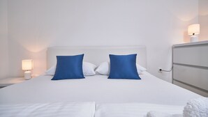 Una cama bien hecha con sábanas blancas y dos almohadas azules, una lámpara de noche y un tocador blanco con espejo.