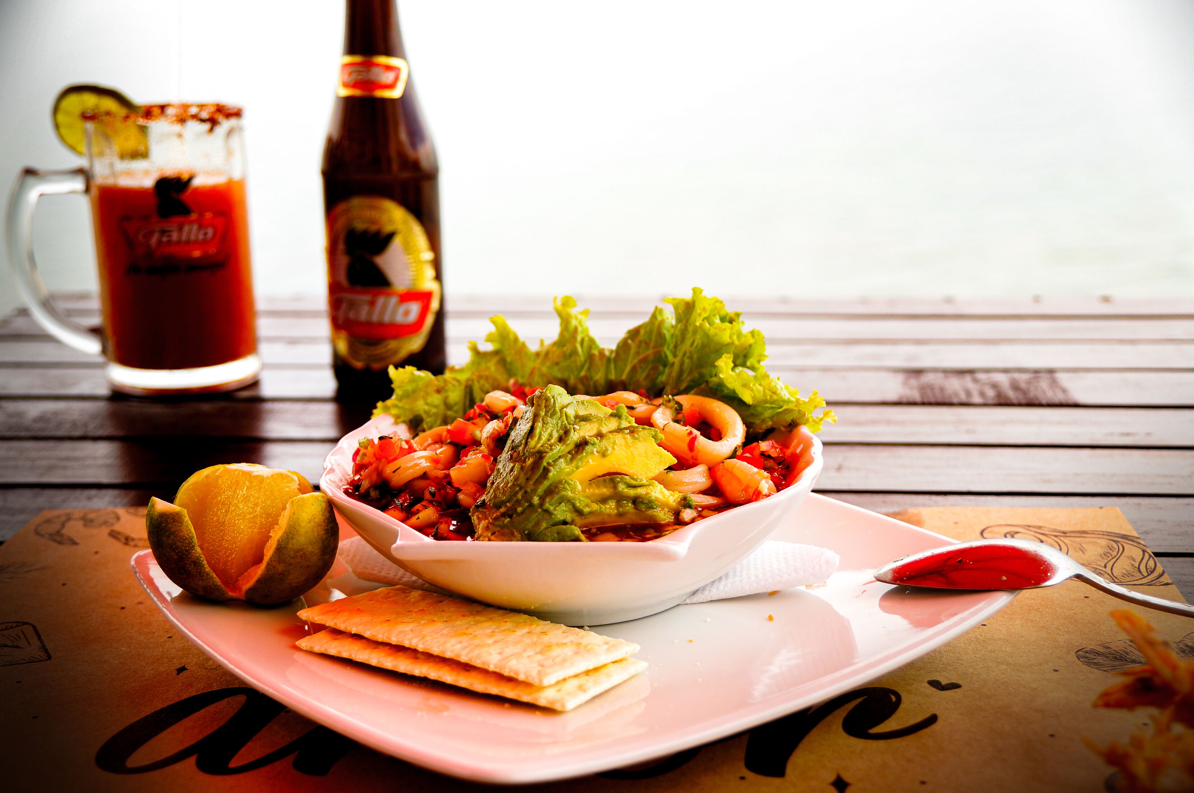 Una comida con un tazón de ensalada, una rodaja de lima, una botella de salsa y un vaso de Bloody Mary sobre una mesa de madera.