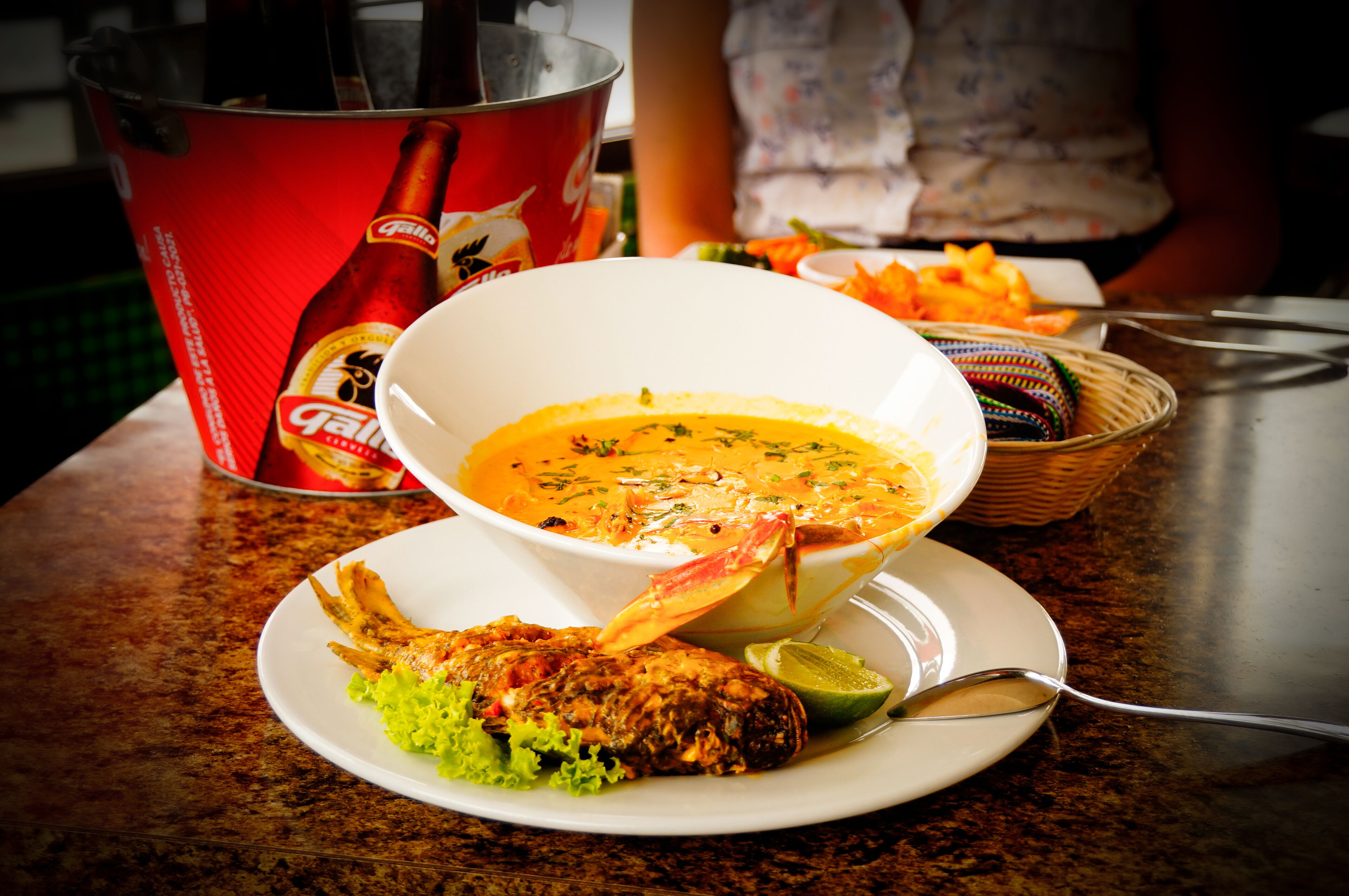 Un tazón de sopa con una cuchara, un plato con pescado y una rodaja de lima sobre una mesa.