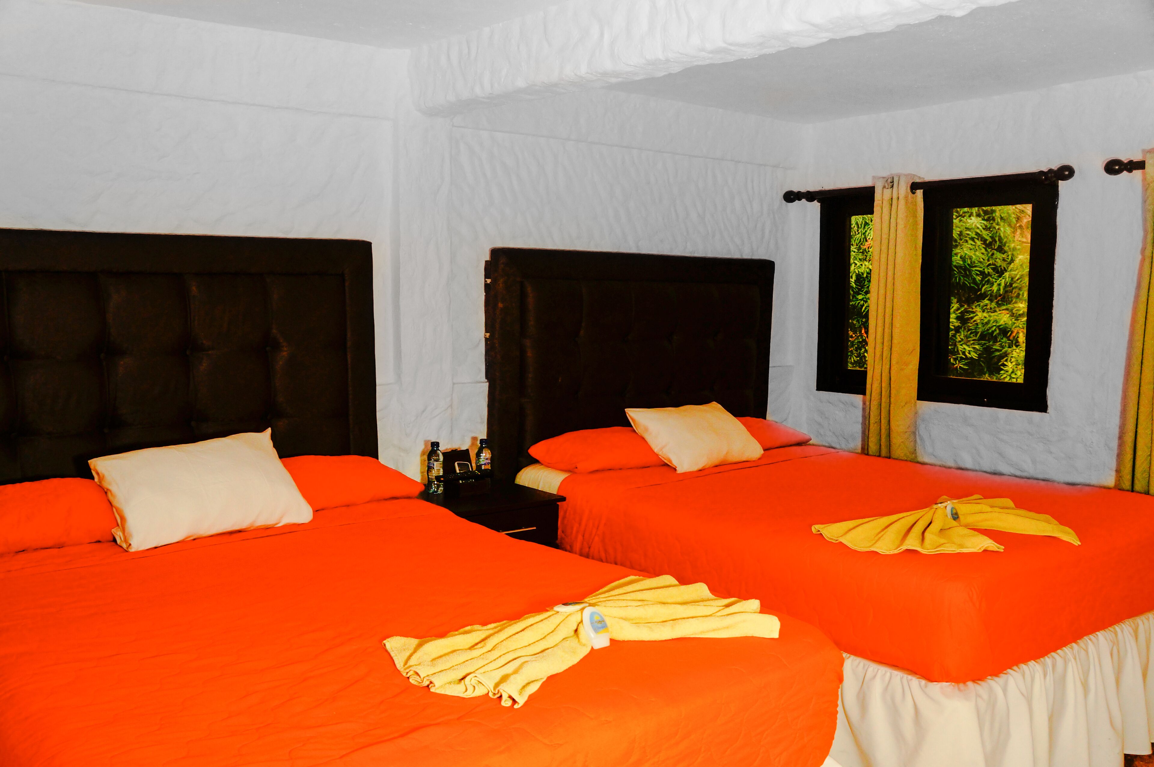 Habitación de hotel con dos camas, cada una con colcha naranja y almohadas blancas. Hay dos mesitas de noche con botellas encima.