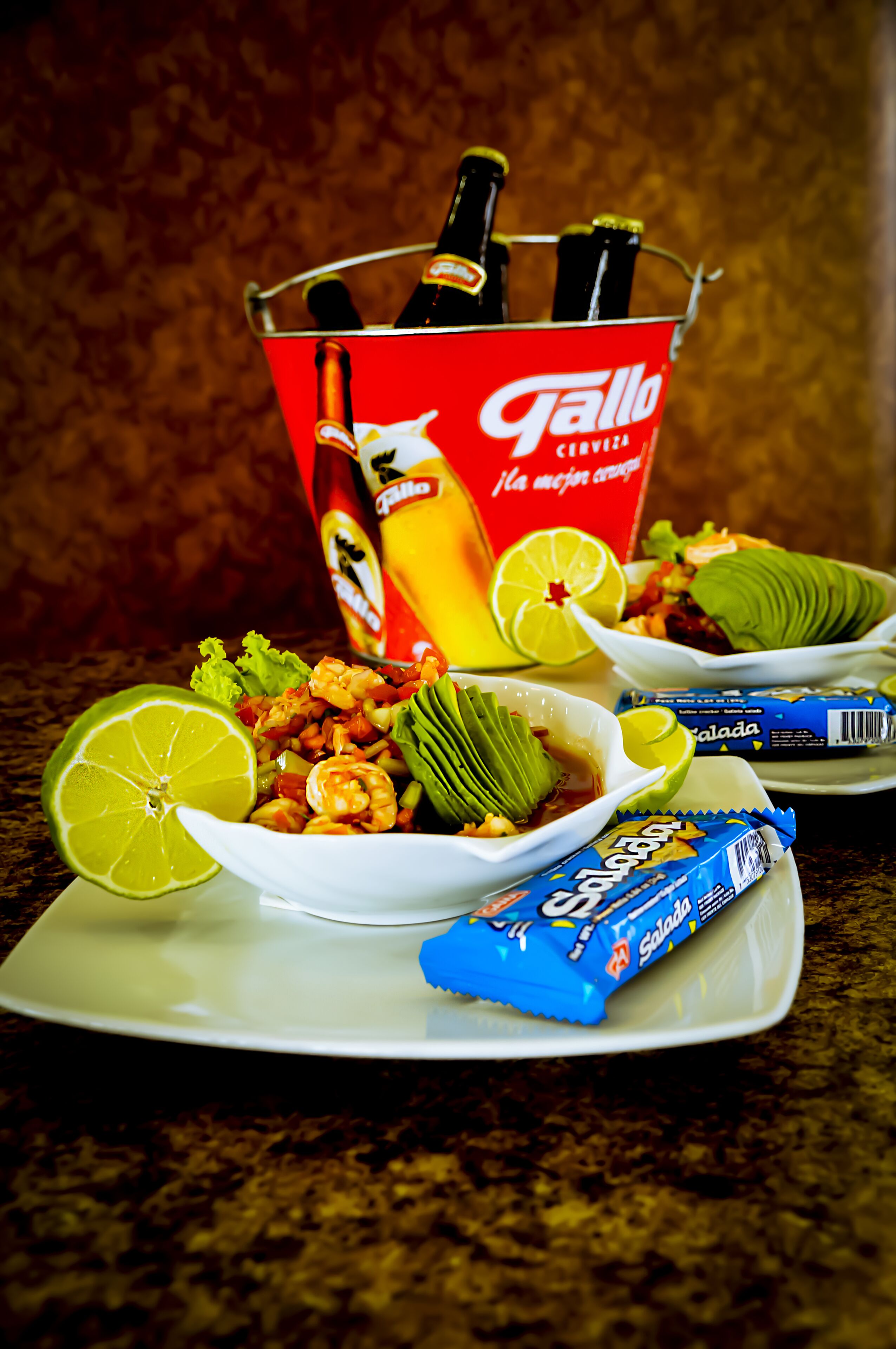 Une table avec un seau réfrigérant pour les bières Gallo, une assiette de salade de crevettes, des quartiers de citron vert et un paquet de vinaigrette pour salade Gallo.
