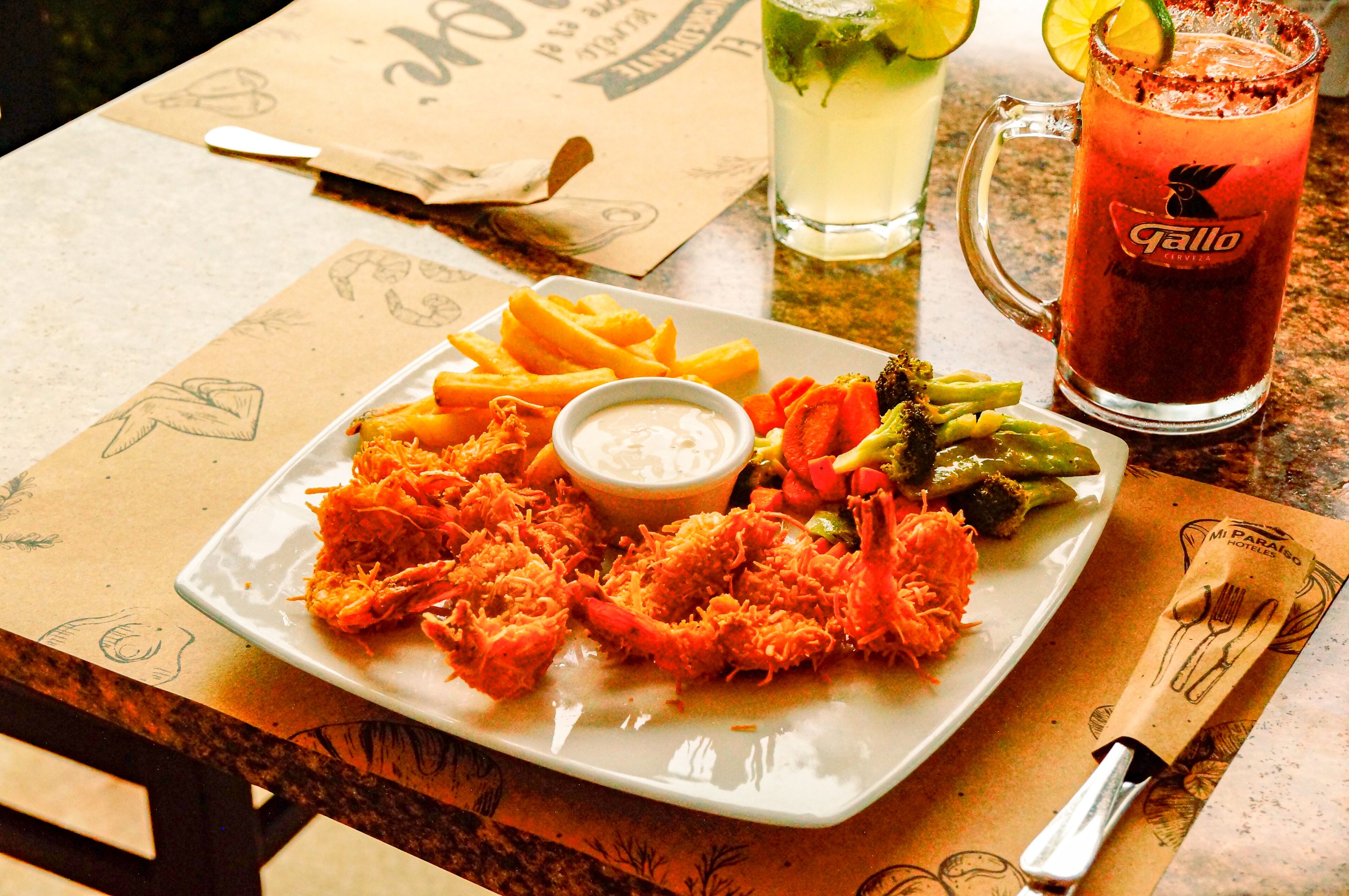 Un plato de mariscos, papas fritas y verduras con salsa para mojar, un vaso de té helado y un cóctel sobre una mesa con un menú y una servilleta.