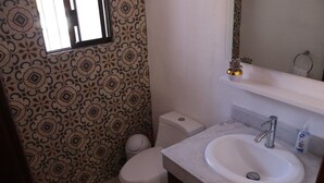 A bathroom with patterned wallpaper, a toilet, a sink with a mirror above it, and a small shelf with a decorative item. - Casas RDM (Mazatlán)