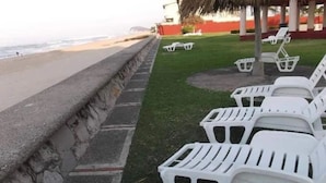 A beachside area with a stone wall, a grassy lawn, and white plastic lounge chairs. - Casas RDM (Mazatlán)