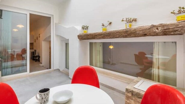 A modern interior with a dining area featuring a white table and red chairs, a glass partition, and a wooden ceiling accent.