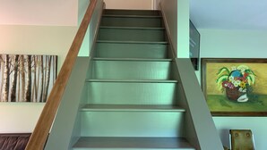Una escalera con barandilla de madera y escalones color turquesa, flanqueada por un cuadro con motivos de bosque a la izquierda y un cuadro floral a la derecha.