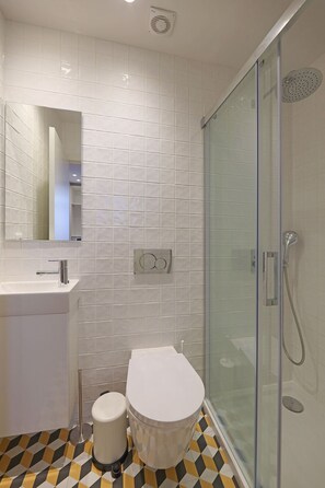 A modern bathroom with white tiles, a glass-enclosed shower, a white toilet, and a sink with a mirror above it. - FLH Porto Downtown GuestHouse (Porto)