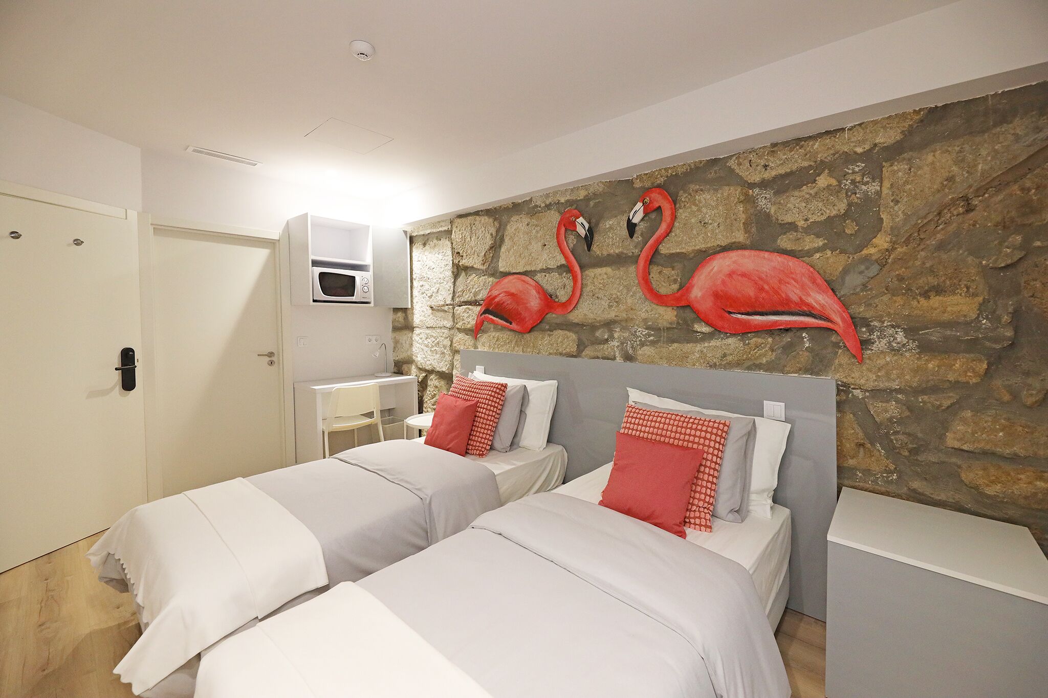 A hotel room with two beds, a stone wall mural, a white door, and a small table with a chair.