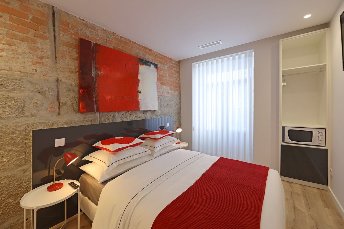 A modern bedroom with a brick wall, a large bed with white and red bedding, a side table with a lamp, a built-in TV, and a walk-in closet.